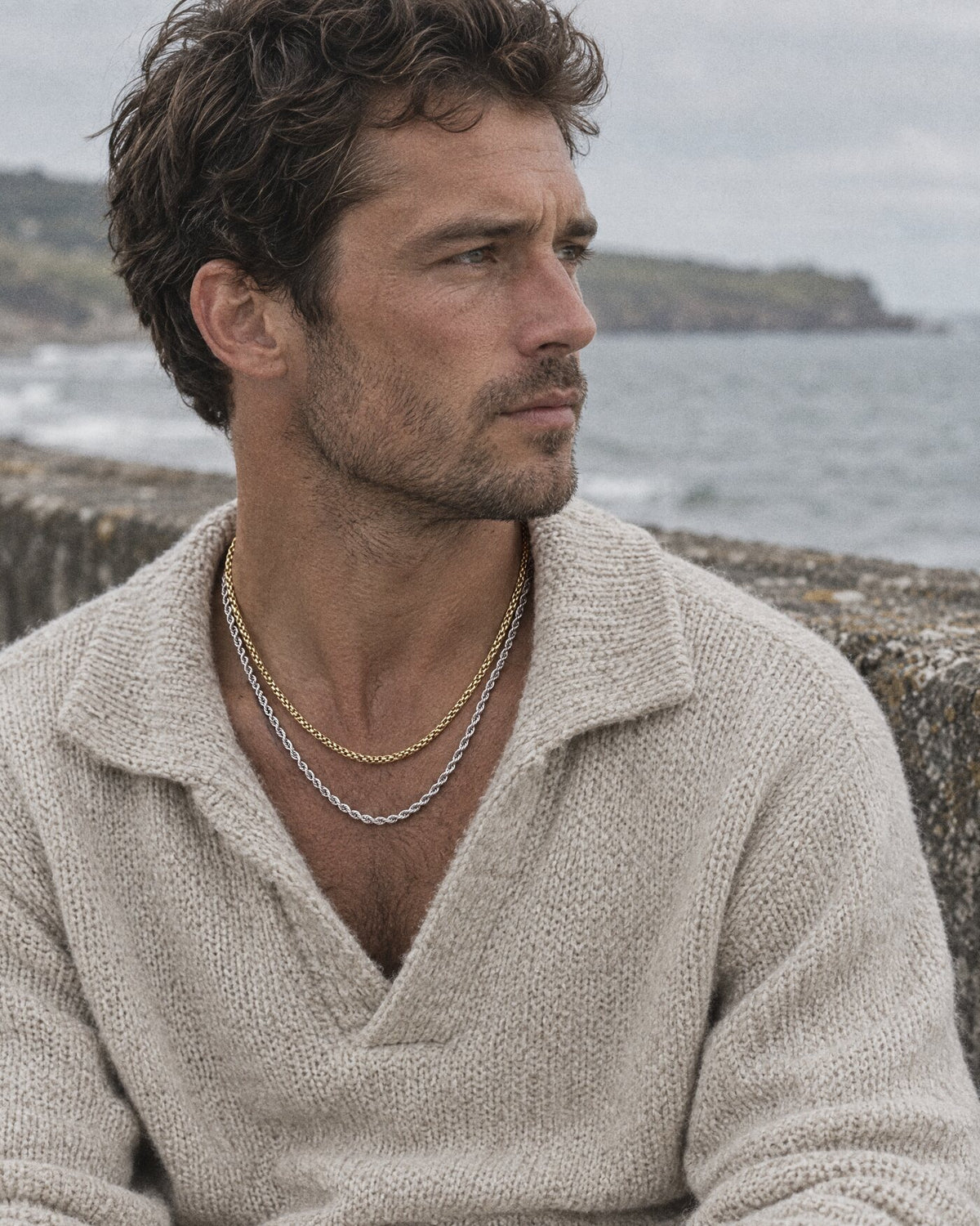 A man in a stone knit on a sea wall layering gold and silver chains