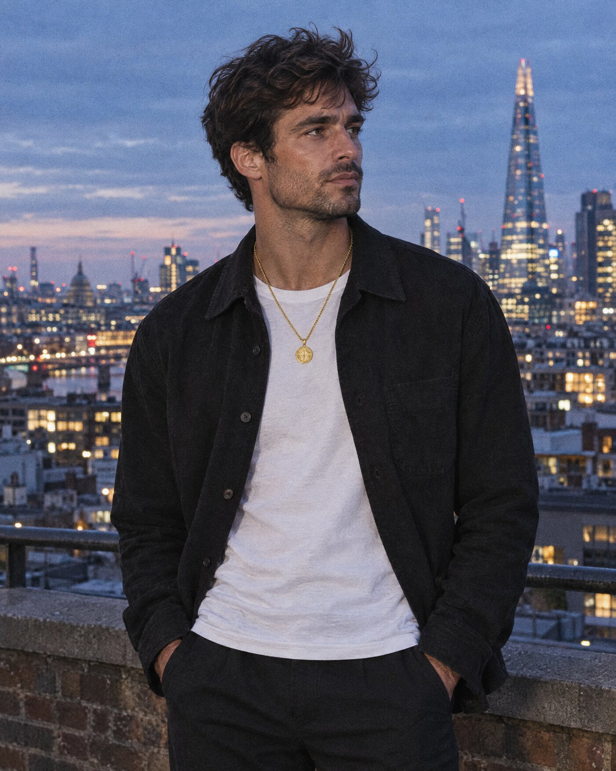 A man on a London rooftop at dusk layering a gold rope chain and a gold compass pendant